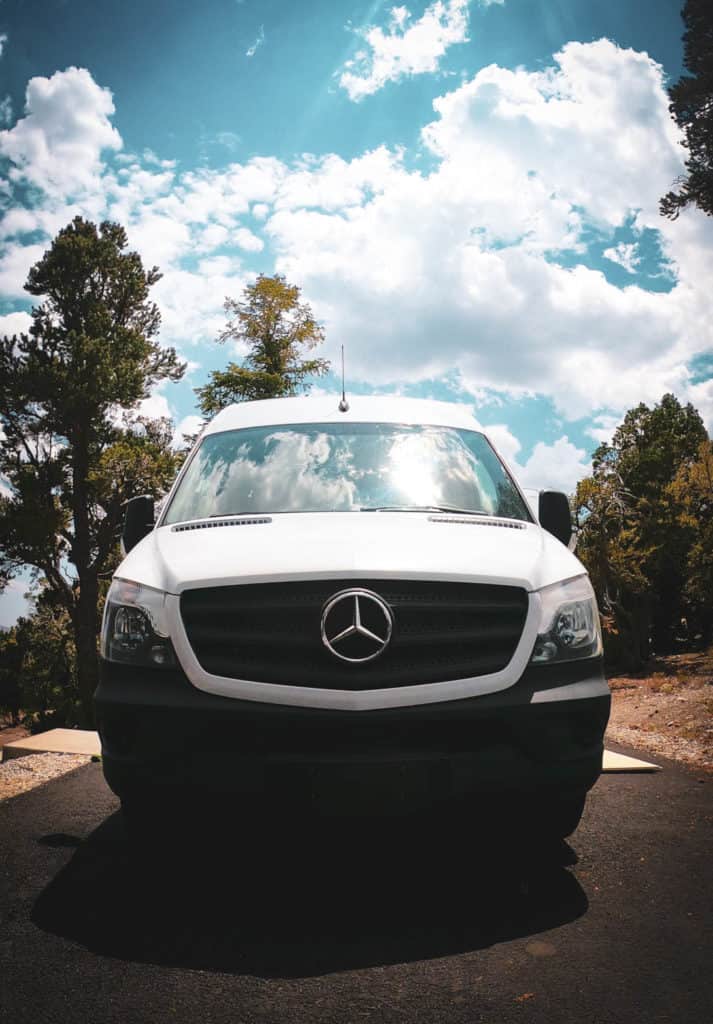 A front view of a white Mercedes Benz Sprinter van. It's parked in the woods. The sky is blue with white clouds.