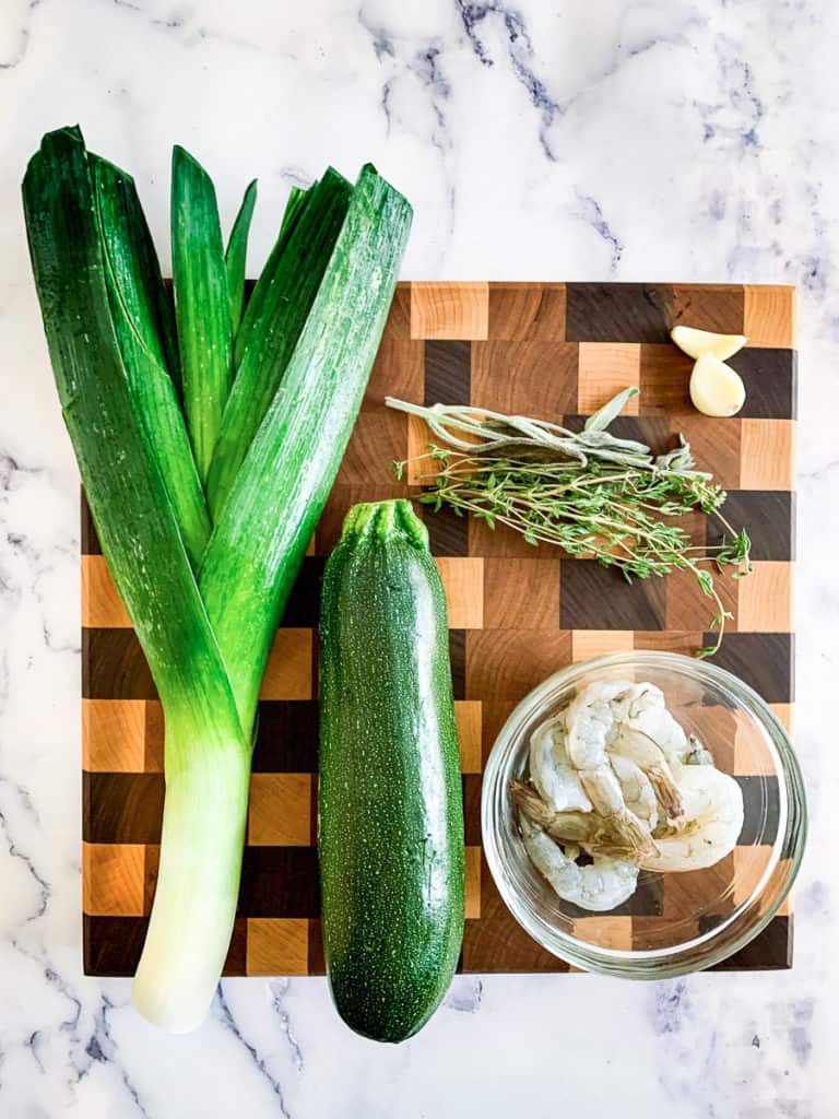 A marble table with a brown cutting board. Leeks, herbs, zucchini, garlic, and shrimp are on the cutting board.