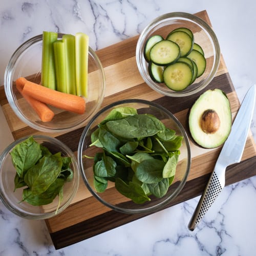 Carrots, celery, basil, spinach, cucumbers, and avodaco on a cutting board with a knife