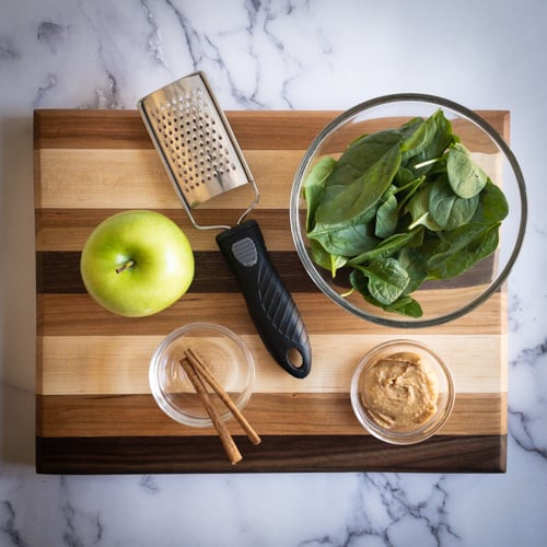 Green apple, spinach, peanut butter, and cinnamon sticks on a cutting board